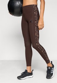 Καφέ κολάν με λεπτομέρεια λεοπάρ στο πλάι, με λογότυπο Adidas. Συνδυάζεται με μαύρα αθλητικά παπούτσια, που παρουσιάζουν κομψό, εφαρμοστό σχεδιασμό.