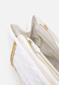 Sac à main blanc matelassé avec un accent en chaîne dorée et plusieurs compartiments zippés. L'intérieur comprend une petite pochette zippée. Texture lisse.