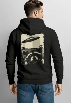 Schwarzer Hoodie mit einem grafischen Druck eines Waschbären und einem UFO, vor einem strukturierten, vintage-inspirierten Hintergrund in Creme- und Schwarztönen.