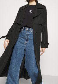 Persona vestida con un largo abrigo negro sobre una camiseta negra de Calvin Klein y jeans anchos de mezclilla azules, de pie contra un fondo sencillo.