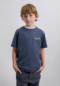 Ragazzo giovane con capelli castano chiaro e lentiggini, indossa una maglietta blu navy con il logo "The Scalpers" e pantaloncini scuri, mani nelle tasche.