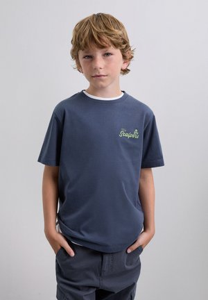 Young boy with light brown hair and freckles wearing a navy blue t-shirt with "The Scalpers" logo and dark shorts, hands in pockets.