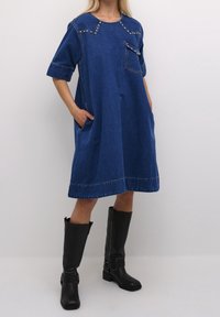 Blå denimklänning med korta ärmar, framficka och silvriga nitar längs axlarna. Kombination med knähöga svarta läderstövlar.