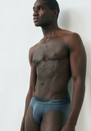 Muskuløs mand i grå Calvin Klein underbukser, der står foran en enkel hvid væg og kigger til siden.