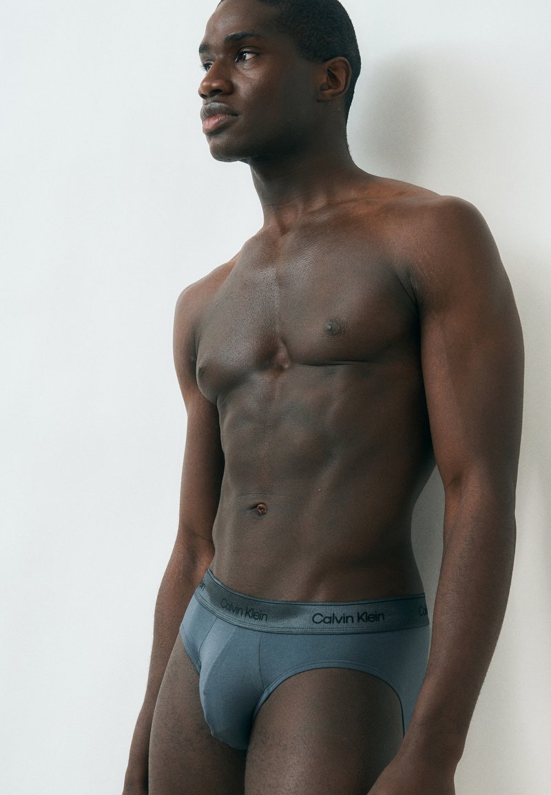 Izmos férfi szürke Calvin Klein alsónadrágban, egyszerű fehér falnak dőlve, oldalra nézve.