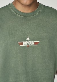 Zelená bavlněná mikina s kulatým výstřihem, zdobená logem "TOP GUN" a grafikou letadla v bílých a červených akcentových barvách. Materiál vypadá jemně.