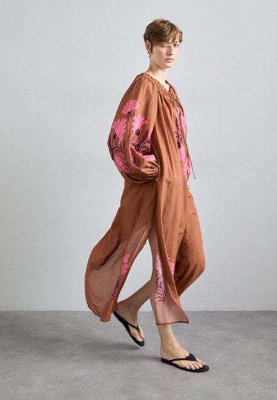 Bruine, lichte, lange mouw jurk met roze bloemenprints, met een losse pasvorm en zijsplitten. Gemaakt in combinatie met zwarte teenslippers.