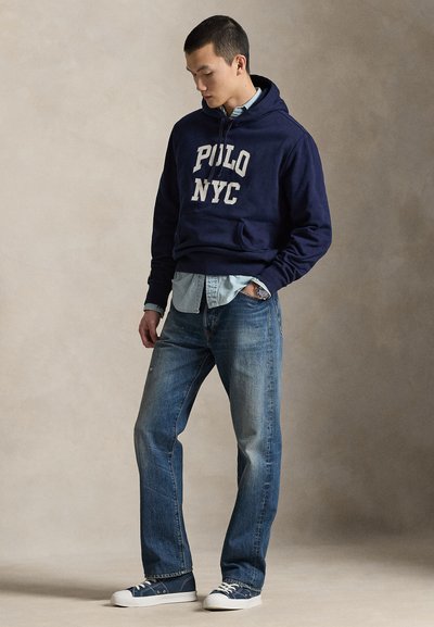 Μπλε σκούρα φούτερ με κουκούλα με τη φράση "POLO NYC" σε λευκά γράμματα, συνδυασμένο με ανοιχτό μπλε τζιν πουκάμισο και ξεθωριασμένο μπλε τζιν παντελόνι. Λευκά αθλητικά παπούτσια ολοκληρώνουν την εμφάνιση.