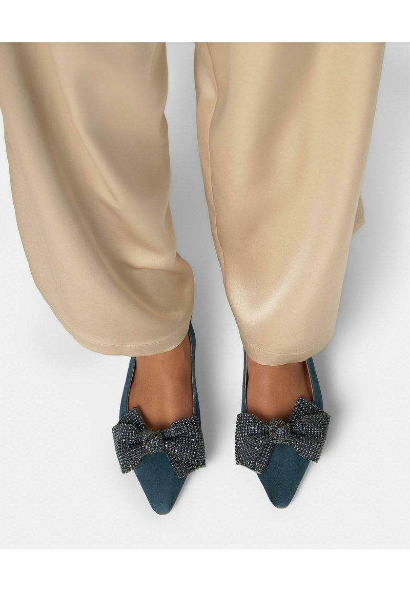 Füße in türkisfarbenen spitzen Ballerinas, verziert mit großen schwarzen und silbernen Perlen-Schleifen, kombiniert mit beigen Hosen mit weitem Bein.