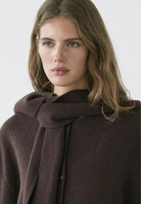 Jeune femme aux cheveux châtain clair ondulés portant une écharpe en tricot marron foncé drapée sur un pull boutonné assorti, fond neutre.