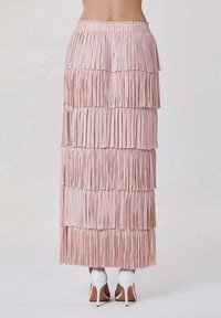 Jupe longue à franges de couleur rose pâle avec une texture en couches, taille élastique et design fluide ; portée avec des chaussures à talons hauts blanches.