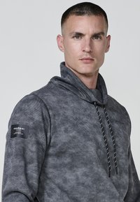 Sudadera gris con una textura sutil, que presenta un cuello alto, cordones con acentos en negro contrastante y un parche de logo en la manga izquierda.