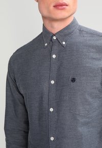 Tmavě modrá košile s límečkem a strukturovanou tkaninou, která má jemný cik-cak vzor, bílé knoflíky a malé tmavé logo na hrudi.