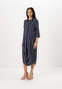 Abito camicia a righe blu navy con bottone sul davanti, maniche a tre quarti e vestibilità comoda, abbinato a sandali con tacco blocco color pesca.