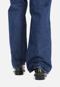 Široké tmavě modré džíny z denimu s texturovaným povrchem, doplněné lesklými tmavě zelenými kotníkovými botami s robustním podpatkem.
