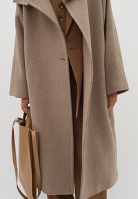 Cappotto lungo beige in lana con maniche ampie, chiusura con un singolo bottone e tasche laterali. Accompagnato da una borsa a tracolla in pelle color sabbia.