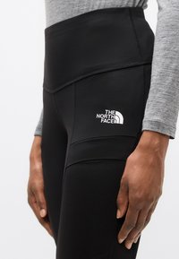 Μαύρα κολάν με ψηλή μέση και λεία υφή, με λευκό λογότυπο, ραφές στις πλευρές και εφαρμοστή σχεδίαση.