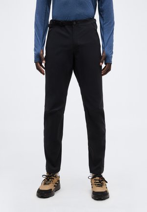 Muž oblečený v čiernych úzkych nohaviciach, modrej košeli s dlhým rukávom a hnedých turistických topánkach, stojaci pred bielym hladkým pozadím.