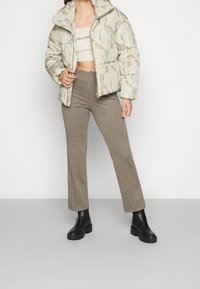 Béžová prošívaná bunda s logovým vzorem, krátký střih, v kombinaci s hnědými kalhotami se vzorem psí zub a černými kotníkovými botami na platformě.