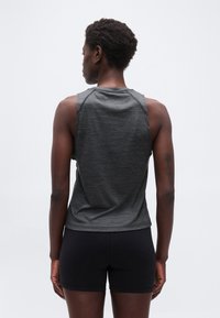 Άτομο που στέκεται με την πλάτη γυρισμένη, φοράει αμάνικη σκούρα γκρι μπλούζα και εφαρμοστά μαύρα σορτς μπροστά σε απλό ανοιχτόφωτο φόντο.