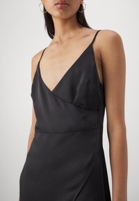 Femme portant une robe noire en satin sans manches avec de fines bretelles et un profond décolleté en V, accessoirisée avec de grandes boucles d'oreilles créoles.