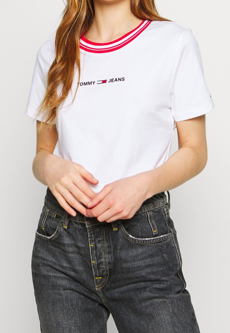 Camiseta blanca con cuello redondo de canalé rojo y texto "TOMMY JEANS". Combinada con jeans oscuros de cintura alta con detalle de costura amarilla.