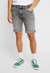 Pantalones cortos de mezclilla gris con dobladillos enrollados, que presentan un acabado desgastado, diseño de cinco bolsillos y cierre frontal con botón. Llevados con zapatillas verdes.
