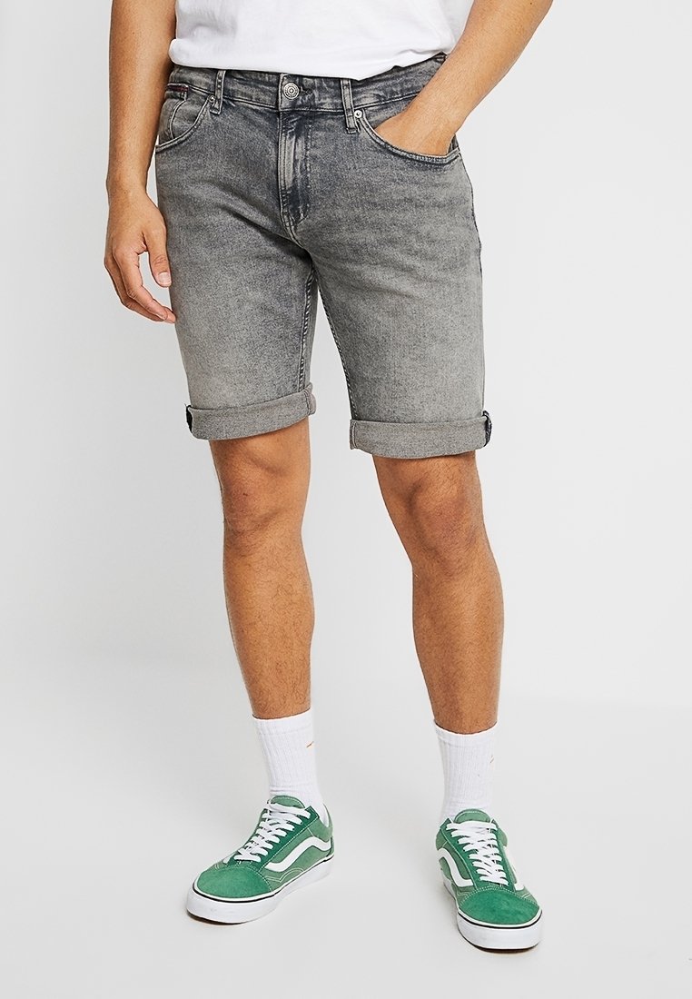 Pantalones cortos de mezclilla gris con dobladillos enrollados, que presentan un acabado desgastado, diseño de cinco bolsillos y cierre frontal con botón. Llevados con zapatillas verdes.