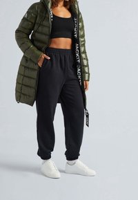 Olivově zelená prošívaná bunda, černý krátký top, černé tepláky s elastickými manžetami a bílé tenisky s nápisem "JACKIT" na boku.