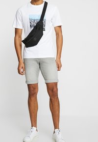 Černá crossbody taška, bílý tričko s grafickým motivem hor, šedé krátké kalhoty, bílé tenisky. Jednoduchý, neformální outfit s funkčními doplňky.