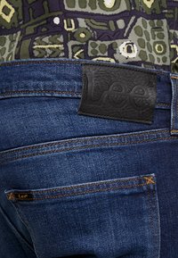 Blå denimjeans med kontrasterande orange sömmar, med en svart läderlogotypetikett med texten "Lee" på midjebandet.