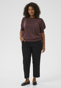 Woman with curly hair wearing a sparkly plum short-sleeve top, black cropped pants, and black ballet flats standing with one hand in pocket.