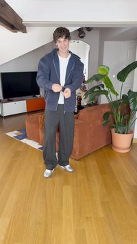 Marineblå hettejakke med glidelås over en hvit skjorte, parret med grå brede bukser og sølvfargede sneakers. Bakgrunnen har en plante og møbler.