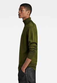 Olivgrüner Rollkragensweater aus einem weichen, gestrickten Stoff. Mit einem gerippten Kragen und Ärmelbündchen, in figurbetonter Silhouette und dezentem Logodetail.