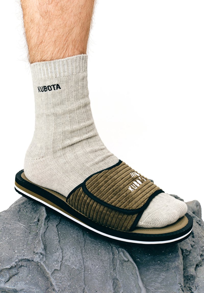 Chausson en velours côtelé brun avec des accents noirs, associé à une chaussette gris clair à côtes portant le texte "KUBOTA". Posé sur une surface gris texturée.
