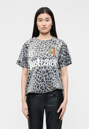 Frau mit langen schwarzen Haaren, die ein graues Leopardenmuster-Shirt mit dem Schriftzug "Just Cavalli" und einem orangefarbenen Sonnenblumen-Aufnäher trägt, kombiniert mit schwarzen Hosen.