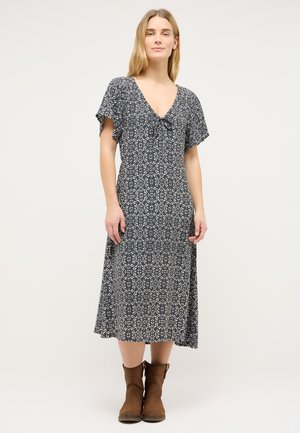 Femme portant une robe midi noire à motif floral blanc, manches courtes, col en V, et bottines marron, debout devant un fond uni.