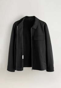 Veste noire avec un col, fermeture éclair intégrale, une poche poitrine et des poches latérales; tissu lisse avec un design minimaliste.