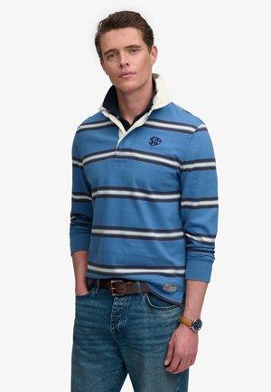 Młody mężczyzna ubrany w niebieską koszulkę rugby w paski z białym kołnierzykiem, niebieskie dżinsy, brązowy pasek i zegarek na rękę, pozuje z rękami w kieszeniach.