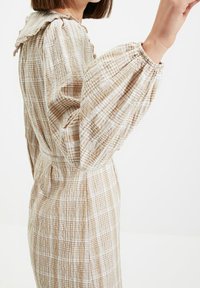 Femme portant une robe à carreaux beige et blanche avec de longues manches bouffantes et un col froncé, vue de côté sur fond blanc.