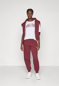 Conjunto de athleisure burdeos que incluye un chaleco sin mangas, una camiseta blanca con el gráfico "PUMA CREW" y joggers a juego. Zapatillas blancas completan el look.