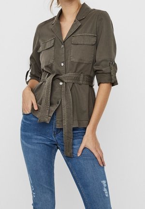 Vrouw draagt een olijfgroene jas met opgestroopte mouwen, ceintuur en voorzakken, en blauwe versleten skinny jeans.