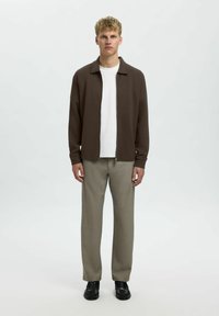 Giacca zip-up marrone sopra una t-shirt bianca, abbinata a pantaloni grigi chiari e texturizzati e scarpe nere. Design semplice, vestibilità standard.