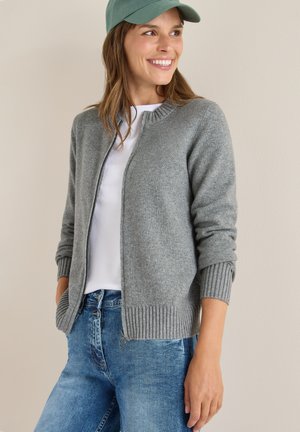 Cárdigan de punto gris con cremallera en el frente, puños y dobladillo acanalados. Combinado con una camiseta blanca y jeans de mezclilla azul. Incluye una gorra de color verde claro.