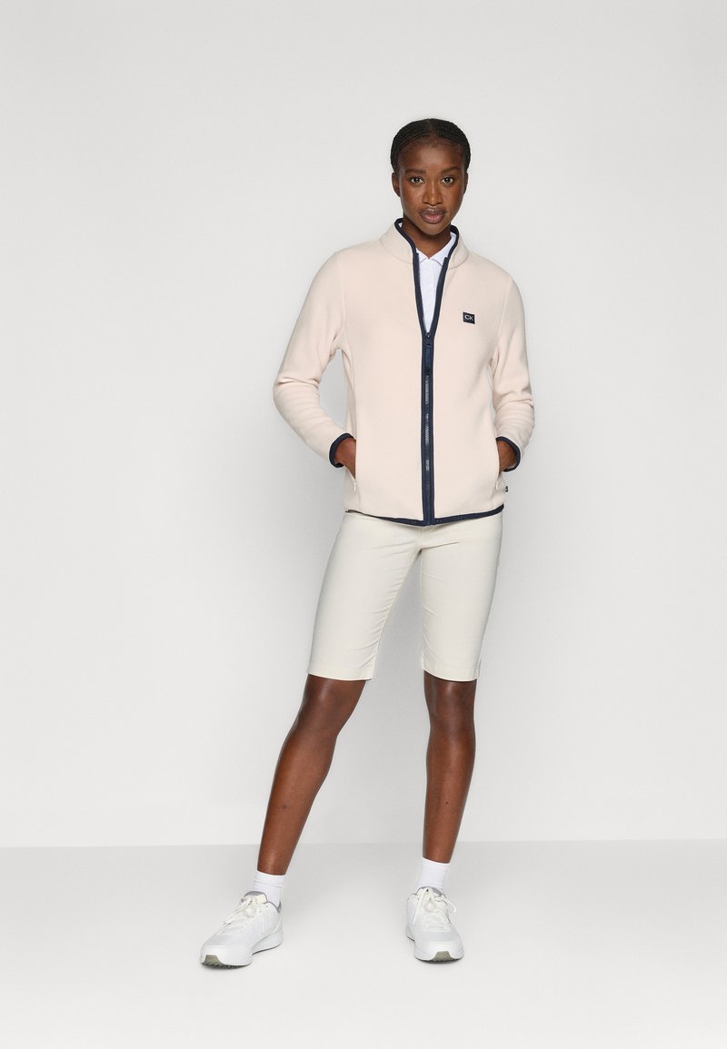 Helle rosa Fleecejacke mit navy Reißverschluss und Akzenten, über einem weißen Polohemd getragen, kombiniert mit cremefarbenen Shorts und weißen Sneakers.