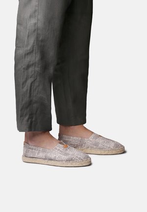 Personne portant un pantalon gris coupé et des chaussures sans lacets avec des motifs géométriques beiges et des semelles tressées, debout sur un fond blanc.