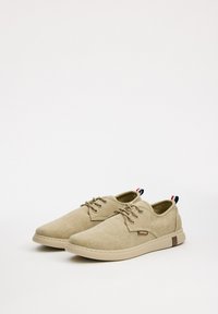 Zapatos de lona beige con puntera redonda, diseño de cordones y acentos marrones en contraste. Presenta una pequeña etiqueta en el costado y un lazo en el talón multicolor.