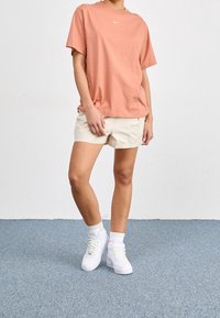 Jasnoróżowy t-shirt z okrągłym dekoltem i białym logo, w połączeniu z beżowymi szortami i białymi trampkami. Materiał bawełniany, casualowy krój.