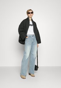 Black quilted oversized jacket, white t-shirt with bold lettering, light blue wide-leg jeans, beige heeled boots, sleek black sunglasses.