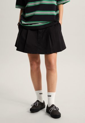 Osoba ubrana w czarną plisowaną spódnicę, zielono-czarny pasiasty top, białe skarpetki Vans i czarne sznurowane buty, stojąca z rękami w kieszeniach.